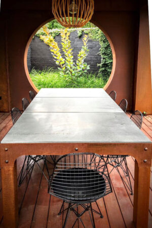 Large concrete-topped dining table with corten pendant light framing circular window view to planted courtyard in Brighton East