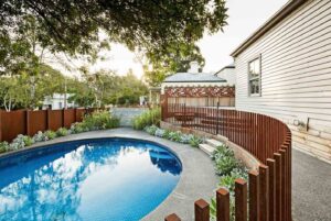 Curved swimming pool with blue mosaic tiles, surrounded by concrete paving and vertical timber slat screening in backyard