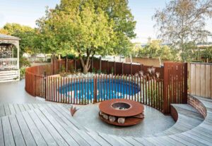 Circular blue pool surrounded by curved corten steel slat fencing, timber decking and central fire pit in Hawthorn backyard