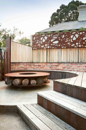 Circular swimming pool with central corten steel fire pit surrounded by curved timber decking and tiered seating areas
