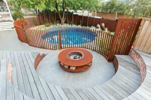 Curved timber decking surrounds corten steel fire pit with circular pool and vertical screening in Hawthorn garden