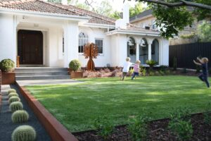 Children playing on expansive lawn with corten steel bottle tree sculpture and structured planting in Malvern East
