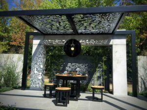Modern perforated steel pergola creating dappled shade over circular dining table and stools in Nagasaki show garden