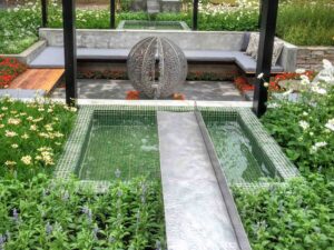 Geometric water feature with circular leaf sculpture and mosaic tiles, surrounded by flowering plants in Nagasaki garden