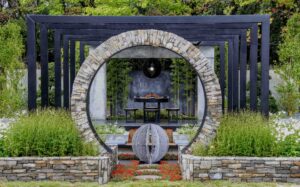 Circular stone moon gate framing outdoor kitchen with steel pergola and sculptural water feature at Nagasaki garden