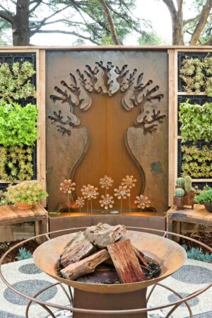 Corten steel tree sculpture backdrop with central fire bowl and flanking vertical garden panels at Melbourne show garden