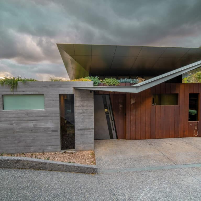 Contemporary coastal home facade with timber cladding, concrete walls and angular roofline set against a dramatic sky.