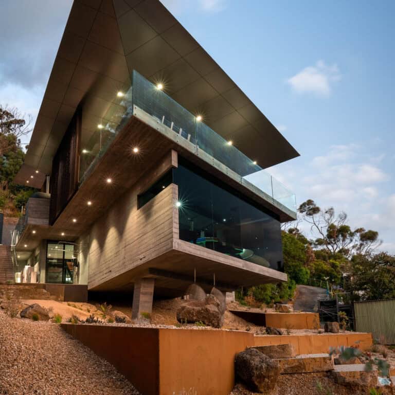 Elevated modern home with corten steel retaining walls and tiered landscaping descending a steep coastal slope.