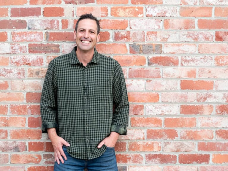 Paal Grant smiling in a green checked shirt, standing against a red brick wall.