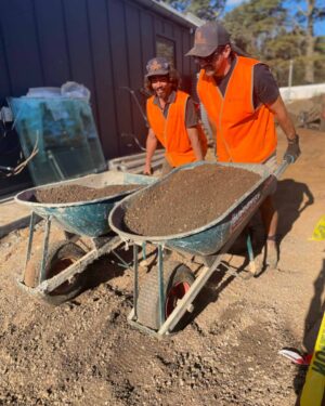 Two workers in high-vis vests preparing wheelbarrows with soil and materials at The Block Daylesford construction site