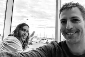 Two people taking a selfie at an airport terminal with aircraft visible through windows, traveling to Nagasaki Japan