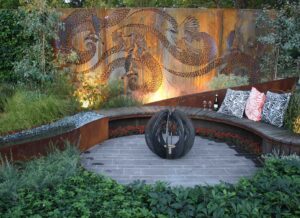 Illuminated Corten steel art screen with wattlebird motifs behind curved stone seating and spherical fire feature in Nagasaki