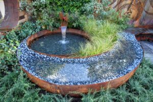 Circular corten steel water feature with central fountain spout and ornamental grass island, surrounded by river pebbles in award-winning Wattlebird Garden, Nagasaki Japan