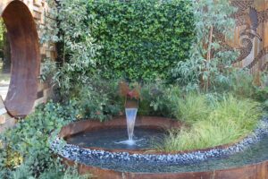 Curved Corten steel water feature with stone spout surrounded by native grasses and decorative river stones in Nagasaki garden