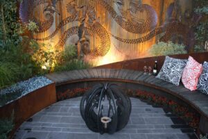 Curved timber bench seating wrapping around central fire feature with decorative metal dragon screen backdrop in Nagasaki