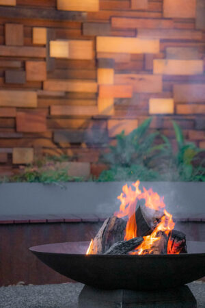 Steel fire bowl with flames burning wood logs, curved corten seating behind, brick wall background in Ballarat garden