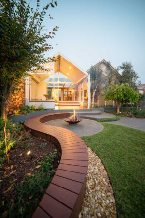 Curved timber deck with central fire pit and corten steel seating overlooking lawn in Ballarat backyard at dusk