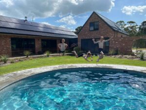 Two children jumping into a circular swimming pool with brick house and solar panels behind at Mount Buninyong garden