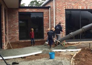 Workers pouring concrete from truck chute while child watches at Mount Buninyong country garden construction site