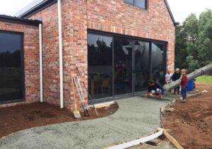 Construction workers pouring concrete for outdoor area at Mount Buninyong country estate with brick house and children watching