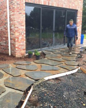 Worker laying irregular flagstone pavers on sand base with brick house and bifold doors visible at Mt Buninyong
