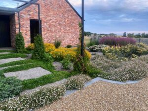 Wide gravel path with stone stepping stones and mixed perennial garden beds beside brick farm buildings, Victoria