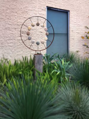 Circular metal ring with arranged stones on timber deck beside palm plants and modern screening