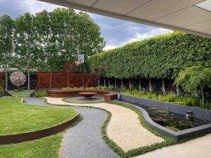 Curved exposed aggregate and gravel pathways wind around circular lawn areas with contemporary water feature and hedge backdrop