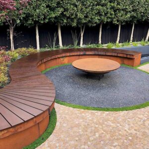 Curved timber bench seating surrounding corten steel fire bowl on gravel surface with landscaped garden backdrop