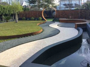 Curved exposed aggregate concrete paths leading to orange steel circular moon gate with water feature and lawn areas