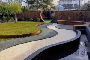 Curved exposed aggregate path leads through circular steel moon gate to lawn area with water feature and timber seating