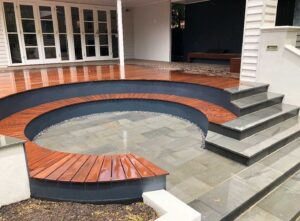 Circular sunken lounge area with curved timber bench seating, bluestone paving, and white rendered steps leading to covered outdoor area