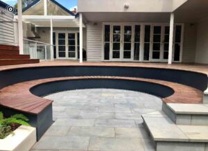 Curved swimming pool with dark corten steel coping and bluestone paving, modern house with French doors in background
