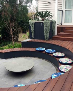 Curved timber deck with built-in seating and colorful patterned cushions around central circular fire pit area