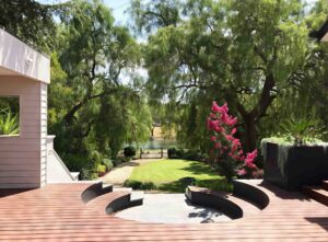 Curved timber deck with built-in circular firepit seating area overlooking landscaped garden with mature trees