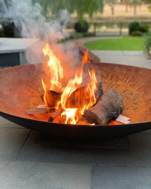 Iron fire bowl with flames burning wood on paved outdoor area with lawn and house in background