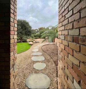 Curved pathway with round concrete stepping stones set in gravel, flanked by brick walls and leading to lawn area