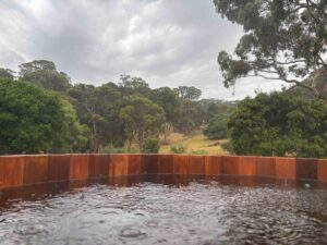 Circular timber spa with water bubbling, overlooking rolling hills and native trees under cloudy sky