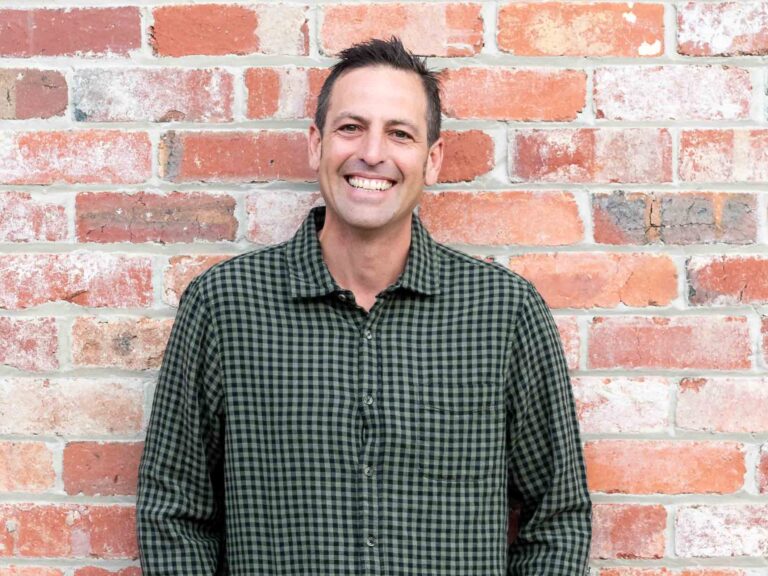Paal Grant smiling in a green checked shirt, standing against a red brick wall.